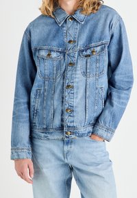 Ljusblå denimjacka med knäppning fram, två bröstfickor och en något kortad passform. Har kontrasterande sömmar och en spetsig krage.