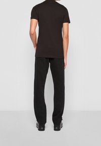 Hombre de pie con la espalda hacia la cámara, vistiendo una camiseta negra de manga corta, pantalones negros rectos y zapatos negros.