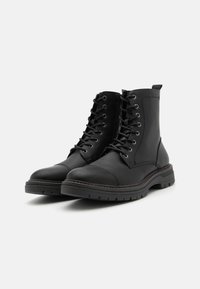 Botas de cuero negro hasta el tobillo con punta redonda, que cuentan con ojales de metal, cordones, suela de goma resistente y un acabado texturizado.