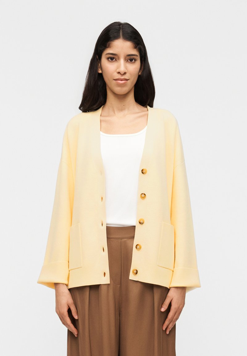 Γυναίκα που φοράει ένα ανοιχτό κίτρινο ζακέτα πάνω από λευκό τοπ και καφέ παντελόνι με φαρδιά πόδια, στέκεται μπροστά σε μια απλή ανοιχτή φωτεινή επιφάνεια.