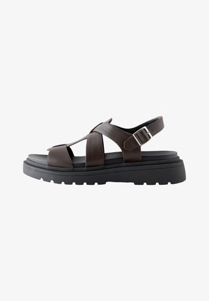 Tmavě hnědá kožená sandal s tlustou černou podrážkou, křížovými pásky a nastavitelnou stříbrnou sponou kolem kotníku.