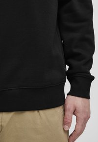 Must духовik musta sweatshirt, millel on soonik ja varrukate ribitekstuur, silmapaistva sileda pinnaga. Käsi hoiab nähtavalt äärest, kõrval beezid pükse.