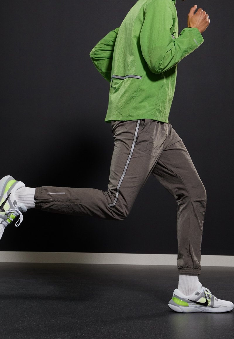 Veste légère verte et pantalon gris avec des rayures réfléchissantes sur les côtés. Chaussures de sport lumineuses avec des accents, chaussettes blanches. Mouvement illustré.