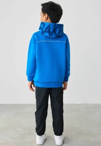 Blauwe sweatshirt met vetgedrukte tekst "BAKER" op de kap, geribbelde manchetten en "Established Nineteen Eighty Eight" op de achterkant. Gepaarde met zwarte cargobroek.