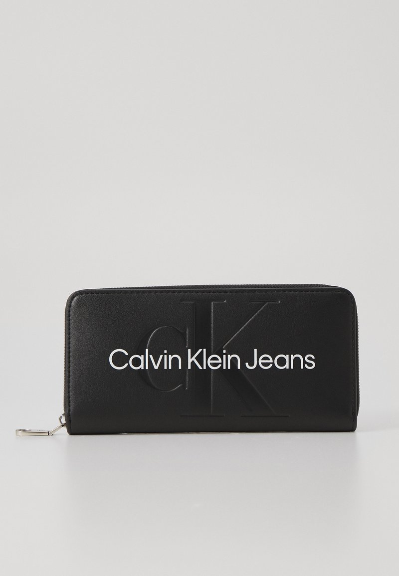 Črna usnjena denarnica z embosiranim logotipom "Calvin Klein Jeans." Ima zadrgo in gladko teksturo. Pravokotne oblike z minimalističnim dizajnom.