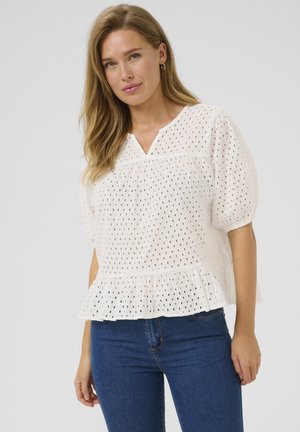 Bela teksturirana bluzica s otvorenimi krogličnimi vzorci, z v-izrezom, kratkimi naborkastimi rokavi in nabranim robom. V kombinaciji z modrimi kavbojkami.