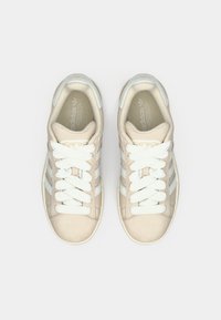 Μπεζ σουέτ αθλητικά παπούτσια με λευκά κορδόνια και τρεις λευκές ρίγες σε κάθε πλευρά. Στρογγυλερή μύτη και επίπεδη ελαστική σόλα. Απλός, μίνιμαλ σχεδιασμός.