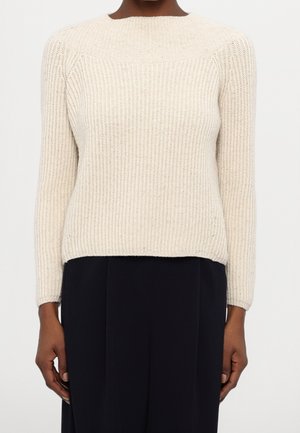 Kremowy, prążkowany sweter dziany z długimi rękawami, noszony na czarnych spodniach przez stojącą osobę na jednolitym tle.