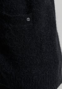 Svart stickad klädesplagg med en texturerad, lockig yta. Har en liten rund logotyp och en framficka.