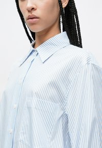 Niebiesko-biała koszula w paski z zapinanym na guziki przodem, kołnierzykiem i kieszenią na lewej piersi. Bawełniana tkanina o gładkiej fakturze i luźnym kroju.