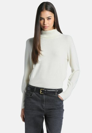 Femme aux cheveux longs et lisses portant un pull à col roulé blanc et un jean foncé avec une ceinture noire sur un fond uni.