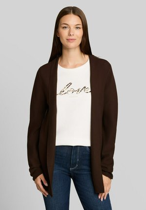 Frau mit glatten braunen Haaren, die eine braune Strickjacke, ein weißes Shirt mit "love" Text und blaue Jeans trägt und vor einem einfarbigen grauen Hintergrund steht.