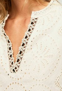 Blouse blanche brodée avec un col en V orné de dentelle, motifs en œillets et tissu texturé avec des motifs floraux.