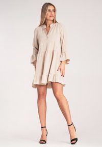 Beżowa sukienka warstwowa z kołnierzykiem, luźny krój, falbaniaste rękawy i dół. Miękka tekstura, średnia długość, odpowiednia na co dzień.