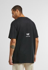 T-shirt en coton noir à coupe décontractée, avec un logo blanc dans le haut du dos et des manches courtes. Texture unie et ourlet standard.