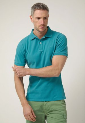 Hombre ajustando la manga de una camiseta polo color azul verdoso, vistiendo pantalones cargo verde claro, de pie frente a un fondo liso y claro.