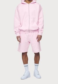 Felpa in cotone rosa chiaro con zip frontale e tasca a marsupio, abbinata a pantaloni corti rosa coordinati. Sneakers bianche e calzini con logo.