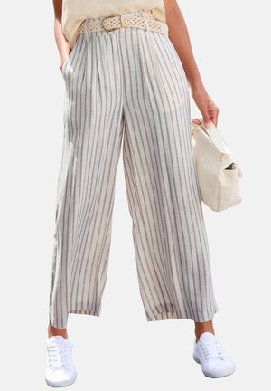 Frau trägt beigefarbene und graue, vertikal gestreifte, weite Hosenbeine, weiße Turnschuhe, beigen Gürtel und hält eine strukturierte beigefarbene Handtasche.