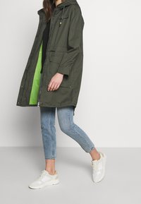 Parka verde oliva con capucha, cierre de cremallera y bolsillos delanteros. Forrada con malla verde brillante, combinada con vaqueros azul claro y zapatillas blancas.