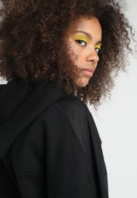 Felpa nera con cappuccio realizzata in un tessuto testurizzato, caratterizzata da una vestibilità rilassata, cordino e un design a strati. Il modello ha capelli ricci e un trucco colorato.