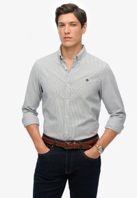 Gestreept overhemd in blauw en wit, gecombineerd met donkere spijkerbroek en een gevlochten bruine riem. Aangekleed met een polshorloge.