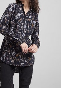 Blommig blus i mörk marinblått tyg, med krage, elastiska ärmslut och en något utsvängd fåll. Designen har flerfärgade blommönster.