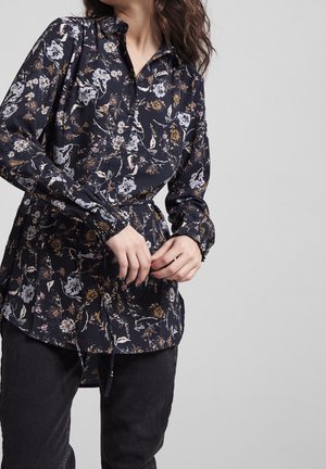 Blouse florale en tissu bleu marine foncé, avec un col, des poignets élastiques et un ourlet légèrement évasé. Le design comprend des motifs floraux multicolores.