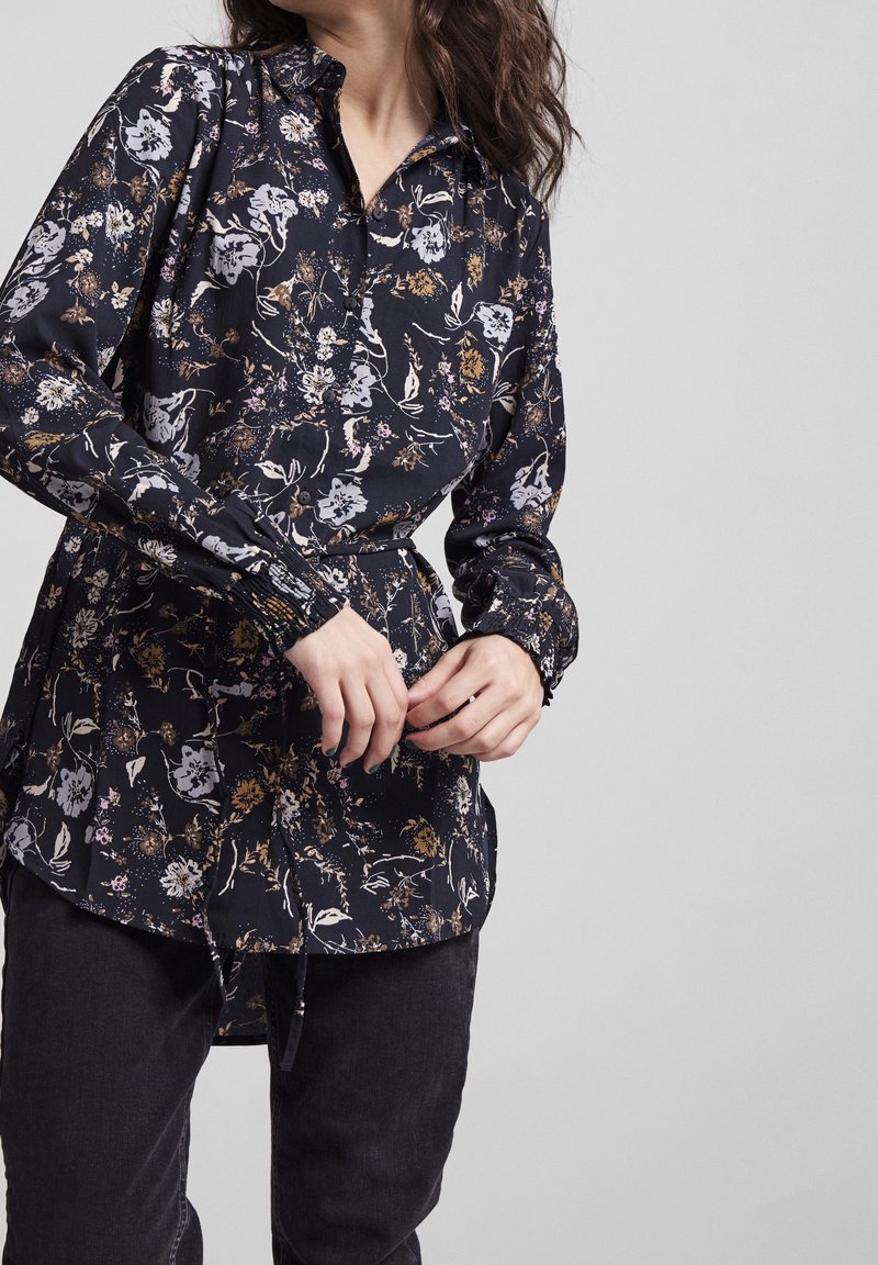 Blommig blus i mörk marinblått tyg, med krage, elastiska ärmslut och en något utsvängd fåll. Designen har flerfärgade blommönster.