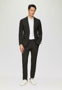 Costume noir avec une veste ajustée et un pantalon slim, associé à une chemise blanche et des baskets blanches. Tissu lisse sans motifs visibles.