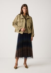 Beige verkürzte Jacke mit zwei Vordertaschen, über einem strukturierten grünen Pullover und einem plissierten Rock mit Farbverläufen in Blau und Braun.