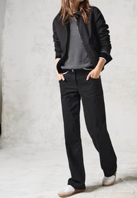 Czarna kurtka na zamek na swetrze w czarno-białe wzory, do tego czarne dżinsy i białe trampki z brązowymi podeszwami.