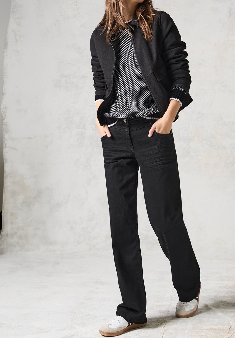 Czarna kurtka na zamek na swetrze w czarno-białe wzory, do tego czarne dżinsy i białe trampki z brązowymi podeszwami.