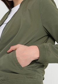 Blouson aviateur vert olive à fermeture éclair frontale, poches latérales et poignets côtelés. Confectionné dans un tissu lisse et léger, avec une silhouette ajustée.