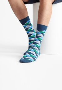 Calcetines con patrón azul y verde con diseño de diamantes, puños acanalados y textura suave, combinados con piernas desnudas sobre una superficie blanca.