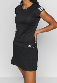 Svart atletisk t-shirt med korta ärmar, slät textur och tre vita ränder på axeln. Adidas-logotyp på den undre sidan.