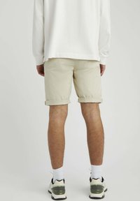 Shorts beiges avec des bords retroussés, portés avec des chaussettes blanches et des chaussures avec des accents verts. La vue de dos montre la texture lisse des shorts.