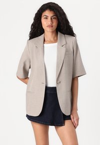 Beige lyhythihainen bleiseri, jossa on yksittäinen nappikiinnitys, toppaukset, kaksi etutaskua ja sileä tekstiili. Yhdistetty valkoiseen puseroon ja farkkuhameeseen.