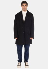 Cappotto di lana nero con colletto a rever e cinque bottoni, dotato di tasche laterali. Indossato sopra una maglietta bianca e abbinato a jeans scuri e sneakers.