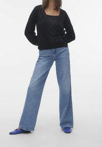 Svart omwrap-tröja i texturerad tyg, tillsammans med ljusblå vida jeans. Blå platta skor fulländar outfiten. Enkel, modern design.