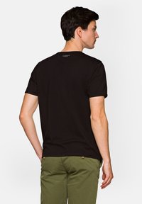 Czarna bawełniana koszulka z krótkim rękawem, okrągłym dekoltem i małym logo na górnej części pleców, pokazana z oliwkowymi spodniami.