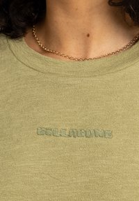 Sweat-shirt vert olive texturé avec un col rond et un logo brodé "BELLABUMB" dans un vert légèrement plus foncé. Collier chaîne visible.