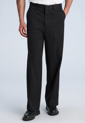Man wearing black formal trousers with pleats, black shoes, and a white shirt, standing with one hand in pocket.