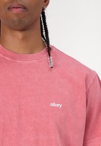 Růžové bavlněné tričko s kulatým výstřihem a krátkými rukávy, na hrudi se malým bílým logem "obey". Měkká textura, uvolněný střih.