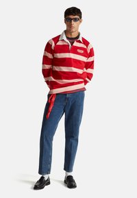 Mężczyzna ubrany w czerwono-różową koszulkę rugby w paski z logo „Stranger Things”, niebieskie dżinsy z czerwoną bandaną, czarne mokasyny, białe skarpetki i okulary przeciwsłoneczne.