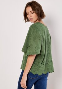 Grünes gesticktes Oberteil mit kurzen Ärmeln, das über florale Ausstanzmuster und einen gewellten Saum verfügt. Kombiniert mit dunklen Jeans.