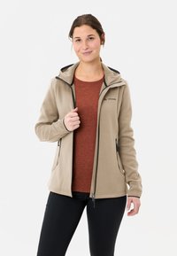 Beige Fleecejacke mit Kapuze, schwarzen Akzenten und Reißverschlusstaschen, über einem rostfarbenen Shirt getragen. Hergestellt aus weichem, strukturiertem Stoff.