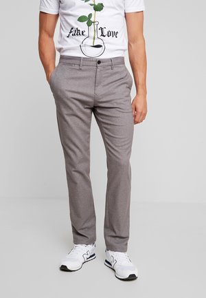 Man draagt grijze, op maat gemaakte broek, wit T-shirt met opdruk 'fake love' en plant ontwerp, en witte sneakers, staat met handen in de zakken.