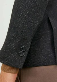 Manica di giacca in tweed nero con una finitura testurizzata, dotata di due bottoni neri e un polsino a coste marrone visibile sotto.