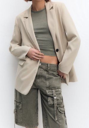Persoon die beige blazer draagt over een korte olijfgroene top met een hoge olijfgroene cargoshort, poserend tegen een effen achtergrond.