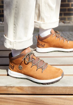 Bruine suède wandelschoenen met grijze accenten, gestructureerde veters en metalen oogjes. Witte tussenzool en logo-detail op de zijkant. Draagt met crème kleurige broeken.